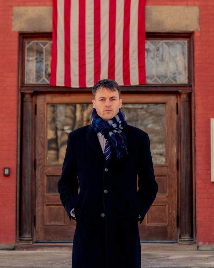 Thomas Plan in a dark coat in front of a brick building; an American flag hangs above the door.
