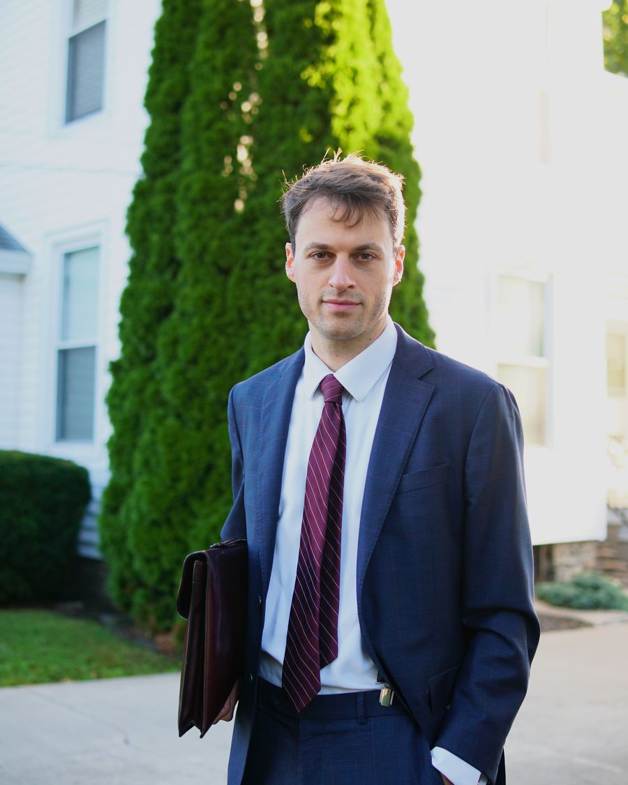 Thomas Plan in a navy suit, a leather briefcase under his arm, standing on a tree-lined street.