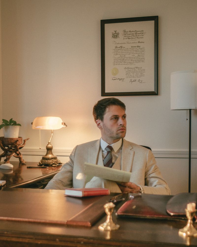 Thomas Plan at his desk, a document in his hand, looking to one side; his New York certificate of admission hangs framed on the wall behind him.